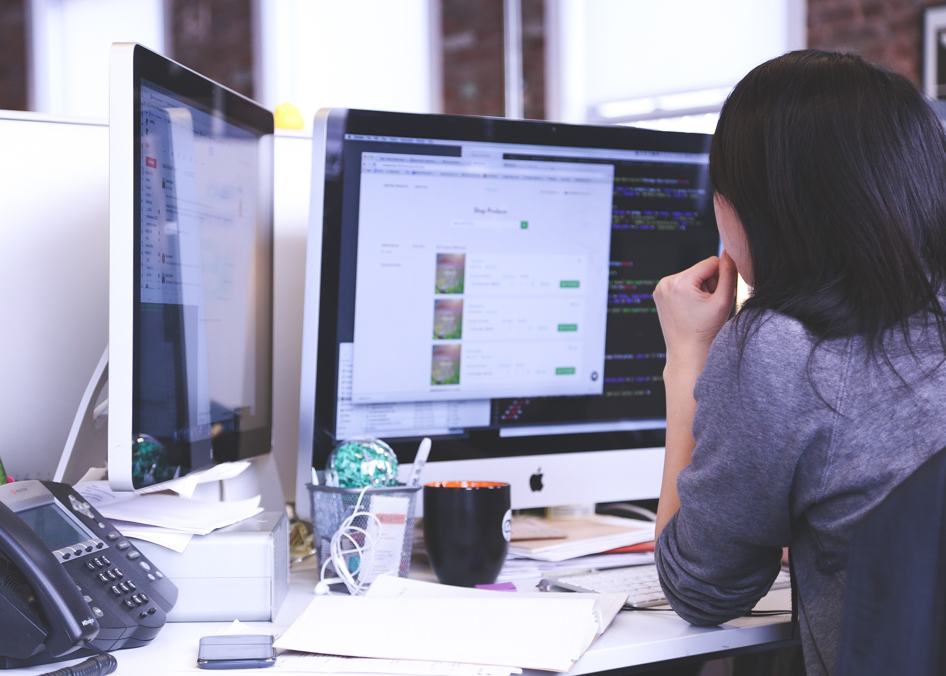 Rückansicht einer Frau, die konzentriert vor zwei Computermonitoren an einem Schreibtisch sitzt, auf dem sich ein Telefon, Tassen und Papiere befinden. Der Hauptbildschirm zeigt eine E-Commerce-Produktseite und dahinter liegenden Programmiercode.