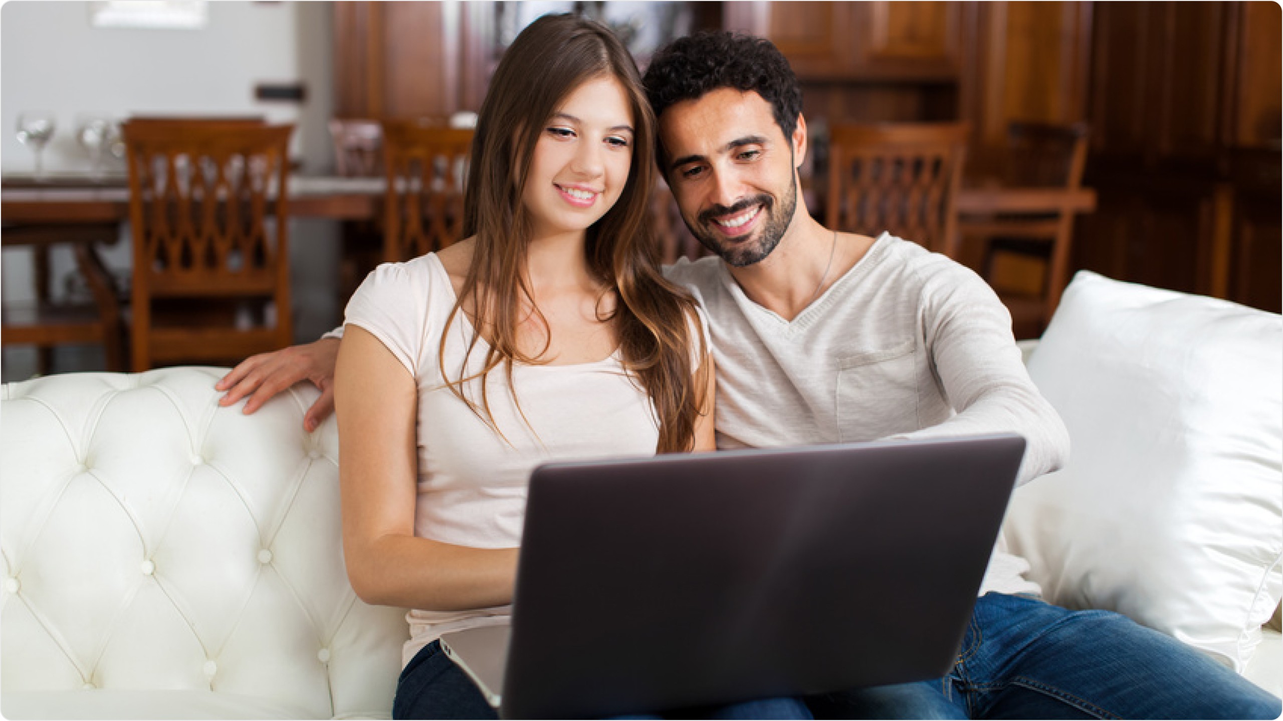 Glückliches Paar sitzt gemeinsam auf weißem Sofa und schaut lächelnd auf einen Laptop in gemütlichem Wohnzimmer mit Holzmöbeln im Hintergrund