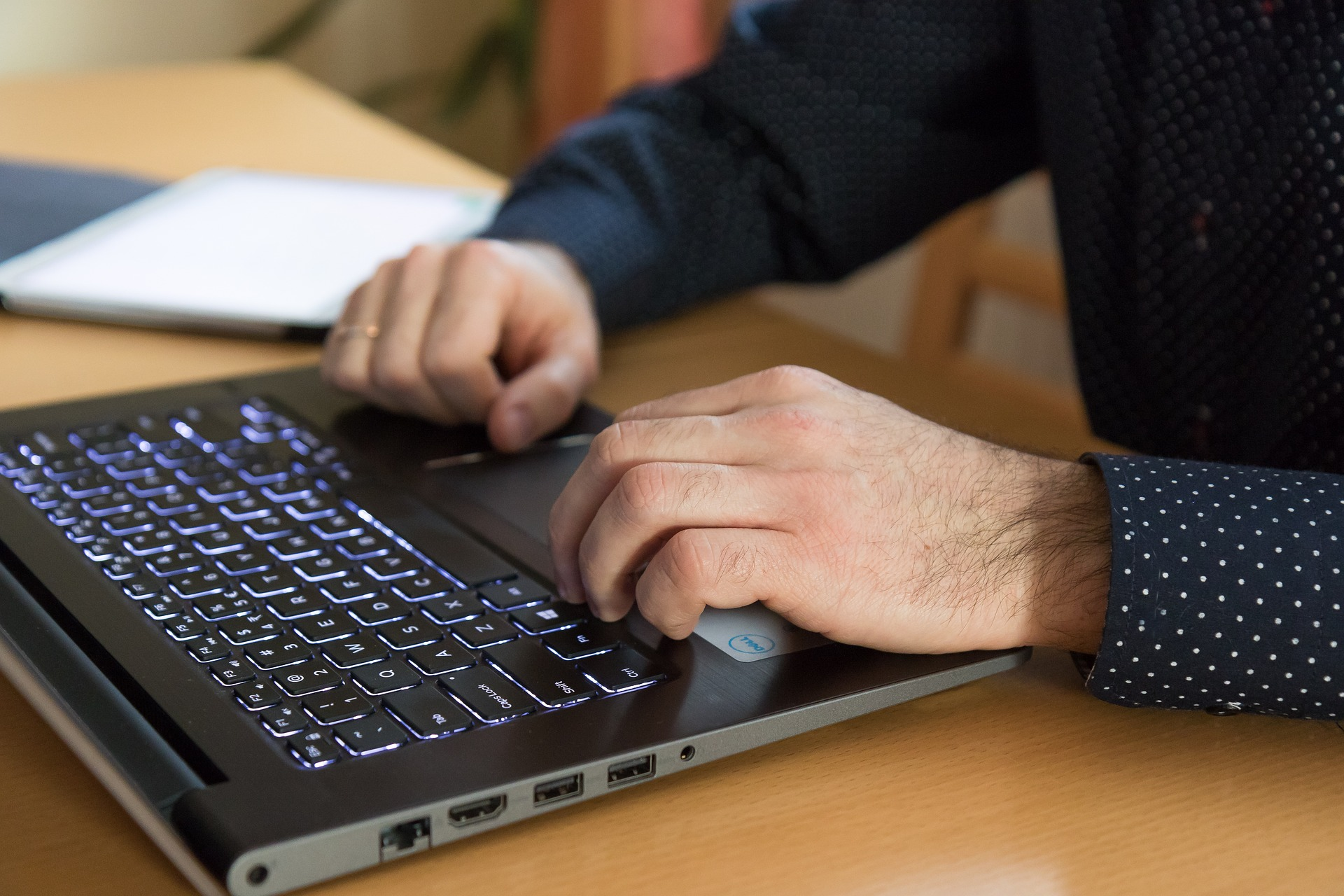Eine Nahaufnahme zeigt eine Person in einem gepunkteten Hemd, die an einem Laptop mit einer blau beleuchteten Tastatur und einem Touchpad arbeitet.
