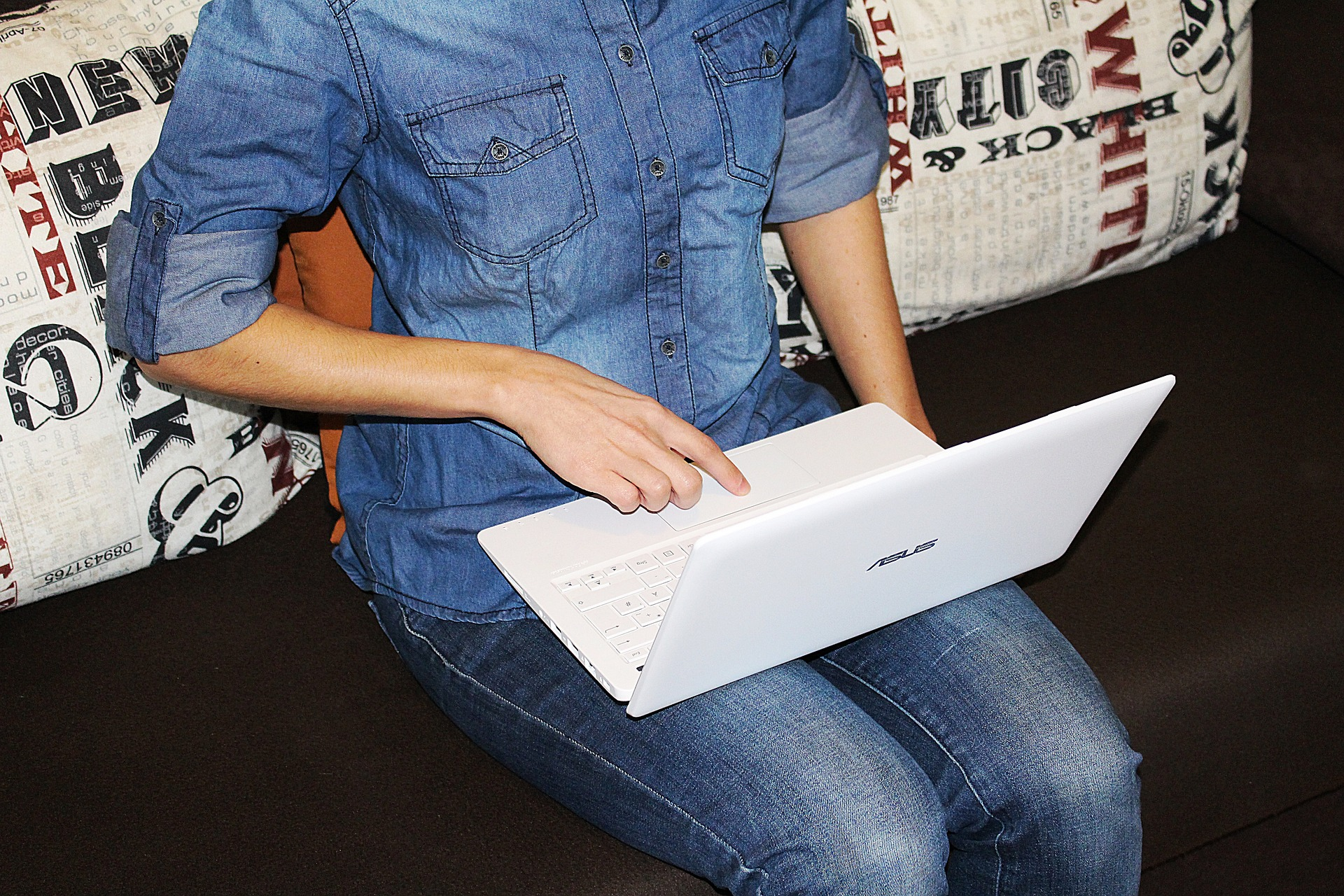 Nahaufnahme einer Person in Jeans und einem Jeanshemd, die auf einem Sofa sitzt und ein weißes ASUS-Laptop auf dem Schoß hält, wobei ein Finger das Touchpad bedient.