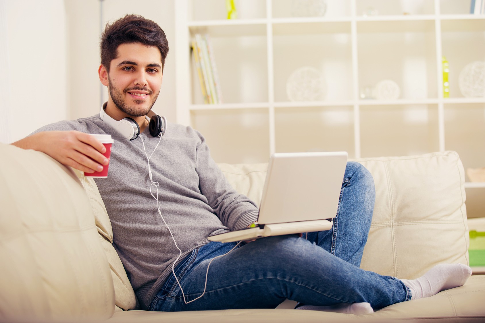 Junger Mann sitzt entspannt auf einem weißen Sofa mit Laptop, hält eine rote Tasse und trägt Kopfhörer um den Hals in einem hellen, modernen Wohnraum.