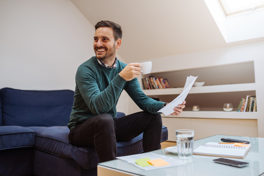 Ein lächelnder Mann in einem grünen Pullover sitzt auf einem dunkelblauen Sofa, hält eine Kaffeetasse und schaut mit Papieren in der Hand nach rechts. Vor ihm steht ein Glastisch mit einem Notizbuch, einem Glas Wasser und einem Smartphone. Im Hintergrund ist ein Bücherregal zu sehen.